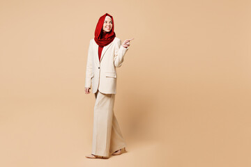 Sticker - Full body side view young Arabian Asian Muslim woman wear red abaya hijab suit clothes point index finger aside on area isolated on plain beige background. UAE middle eastern Islam religious concept.