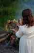 Girl with floral wreath in hands near campfire outdoor, dark natural background. rear view. Magic ceremonial, witchcraft. witch wiccan ritual for Beltane, Midsummer, Litha.