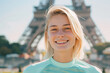 A woman with blonde hair is smiling and standing in front of the Eiffel Tower