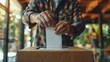 A person's hands inserting a ballot into a voting box or ballot box. The person is wearing a patterned shirt, suggesting a voting or election setting.