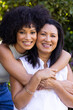 Biracial mother and adult daughter are hugging, smiling outdoors at home in the garden