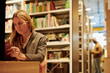 © Clique Images - Serious blond woman sitting in front of camera in modern library and reading book against huge stacks of bookshelves