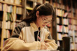 © TrueFrame Collective - Young Asian female student with pen writing down main points of online book while preparing for seminar in reading hall