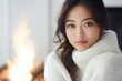 © useful pictures - Beautiful asian american Woman sitting on chair by the fireplace at modern house on nature during winter time. Concept of winter mood and comfort at home. Pretty Girl wearing white sweater