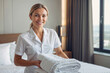 European woman housekeeper in white uniform is holding a stack of white towels and smiling in a luxury hotel room. Service concept
