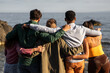 A tight-knit group of friends stands together, gazing out at the sea, sharing a tranquil moment by the ocean, signifying unity and contemplative peace - back view.
