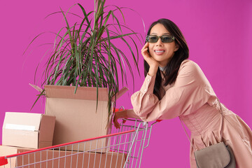 Canvas Print - Beautiful young stylish Asian woman with shopping cart, boxes and houseplant on purple background