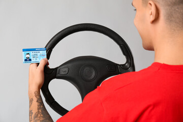 Sticker - Young happy man holding driver license and steering wheel on grey background, back view, closeup