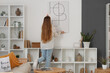© Pixel-Shot - Young woman hanging poster on light wall in living room, back view