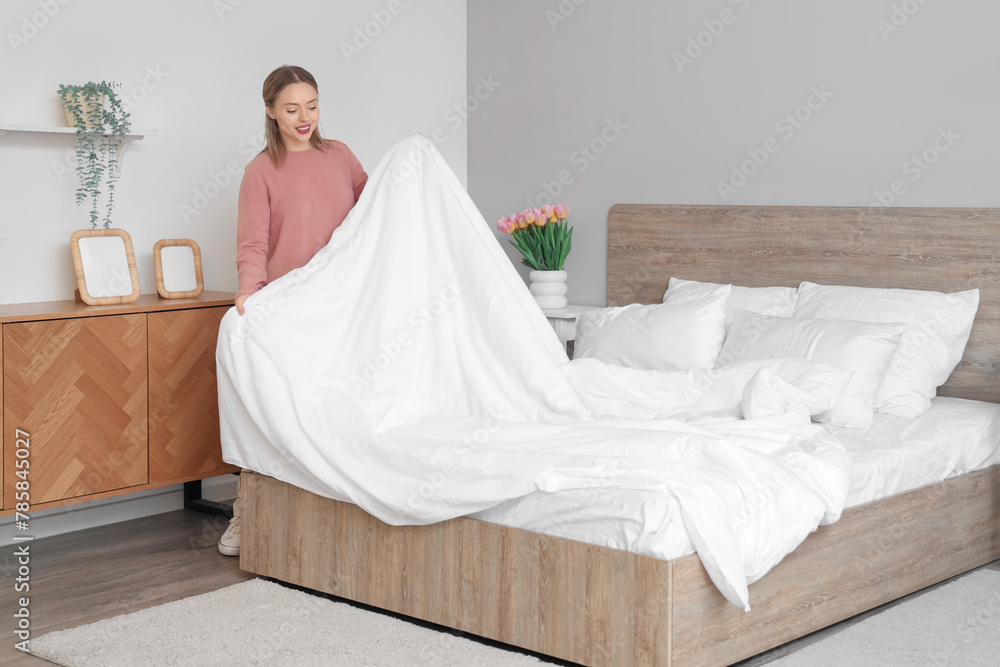 Pretty young woman making bed in light bedroom