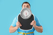 Shocked young man with tape measure around his waist and scales on blue background. Weight loss concept