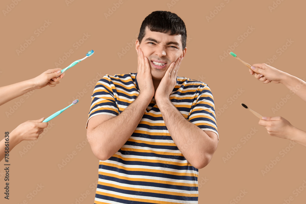 Young man suffering from toothache and hands with brushes on brown background