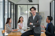 © crizzystudio - Strong young Asian businessman A muscular man smiles at the camera while leaning against his desk, arms crossed happily in the conference room of a modern workplace.