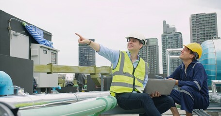 Canvas Print - engineers manager and worker sitting on rooftop review plans on a laptop pointing to something at construction site with high-rise buildings