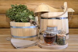 Fototapeta Koty - A glass cup of herbal tea stands on a wooden bench next to a dry birch broom and traditional sauna accessories in the interior of a log bathhouse. 