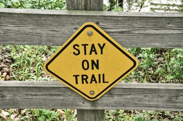 Closeup of a warning yellow sign to stay on trail on a wooden fence