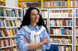 © Liubomir - A cheerful, confident woman with curly hair stands in front of a library bookshelf, wearing glasses and a casual outfit, expressing positivity and intelligence.