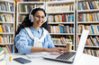© Liubomir - Smiling woman in a library setting using a laptop with headphones. She is surrounded by expansive bookshelves, exemplifying a productive research environment.