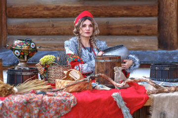 Wall Mural - A beautiful Russian girl in traditional Russian clothes and a kokoshnik stands at a table with various national dishes near a wooden house. Festive farewell to winter.