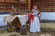 A beautiful Russian girl in traditional Russian clothes and a kokoshnik stands at a table with various national dishes near a wooden house. Festive farewell to winter.