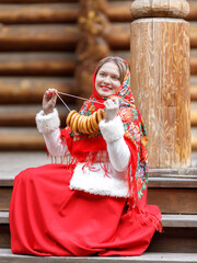 Wall Mural - A beautiful Russian girl in red traditional Russian clothes holds bagels in her hands in the spring near a wooden house. Festive farewell to winter.