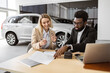 © sofiko14 - Caucasian rich woman customer buyer client in suit sign paper contract document with African salesman choose auto, buying new automobile in car showroom vehicle salon dealership store motor show.