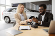 © sofiko14 - Happy Caucasian young woman sitting at table and receives keys of SUV car from African man salesman in suit in bright showroom. Concept of buying new modern vehicle.