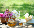 glass teapot, cup with herbal tea, flowers in basket and book on table in garden. Summer nature background. Beautiful rustic composition. reading, relax time. useful calming tea. Tea party outdoor