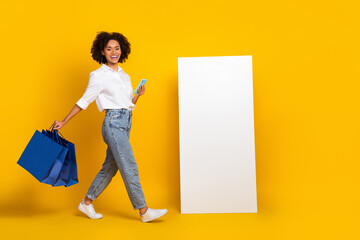 Sticker - Full length body size view of beautiful trendy cheery girl using gadget carrying goods isolated over bright yellow color background