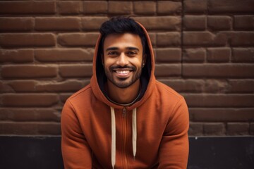 Wall Mural - Portrait of a happy indian man in his 20s sporting a comfortable hoodie while standing against vintage brick wall