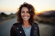 Portrait of a grinning woman in her 40s sporting a stylish varsity jacket in front of vibrant sunset horizon