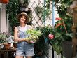 A woman in a straw hat and denim overalls tends to her balcony garden, showing a healthy potted plant