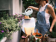 An individual waters plants using a white watering can amid various gardening tools and pots