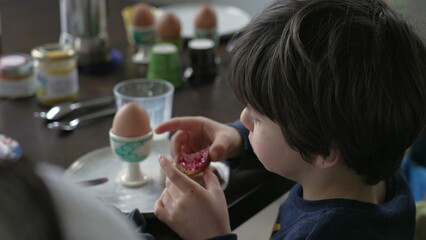 Back of little boy eating toast with jelly seated at breakfast table, close-up of 5 year old boy kid enjoying morning ritual with family, candid and authentic