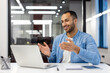 © Tetiana - A smiling young Muslim man sitting in the office at a desk in front of a laptop and talking on a video call