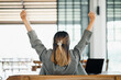 View from behind of a businesswoman with raised arms in victory, celebrating success at her desk with a laptop.