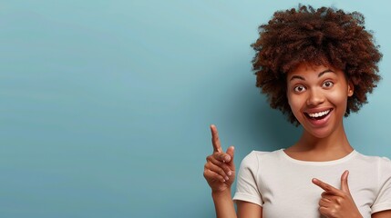 Horizontal shot pleased dark skinned female with Afro haircut, points away with both fore fingers, shows blank space for your promotion, isolated over blue background.. copy space for text.