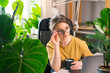 Beautiful young woman drinking coffee at work from home surrounded by indoor plants. Concept of remote work, freelancing, online learning, in the urban jungle.
