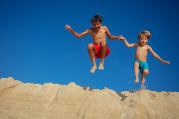 Wall Mural - Pair of cheerful children jump from a sandbank against blue sky