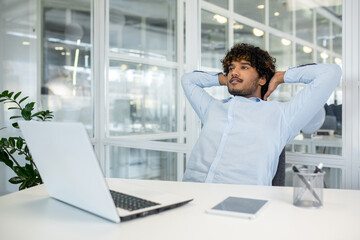 A young man with curly hair sits back, hands behind head, reflecting or taking a break in a well-lit office environment. The scene evokes a sense of calm and contemplation.