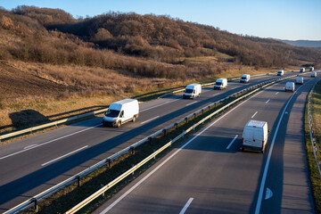 Wall Mural - Eleven white minivans are moving in a convoy on the highway. White delivery van on the highway. White modern delivery small shipment cargo courier van moving fast on motorway road to city urban suburb