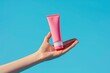 Hand holding a pink tube of cream against a blue background. Suitable for beauty and skincare concepts