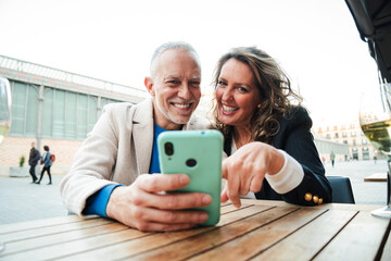 Middle aged couple having fun sharing media with a cellphone. Mature people watching the smartphone on a coffee shop, restaurant or bar. Mid adult friends buying on a market place using a mobile phone