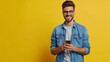 Portrait of a Charming male entrepreneur with hand in pocket smiling and messaging over mobile phone. Happy young man wearing denim shirt and eyeglasses standing isolated on yellow background