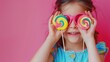 Little sweet tooth. Adorable girl covering her eyes with two colorful lollipops, pink panorama background