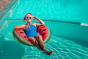 A happy woman in a blue bikini, a red and white Santa hat and sunglasses poses in the pool in an inflatable circle with a watermelon pattern, holding a glass of champagne in her hands.