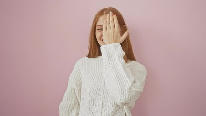 Wall Mural - Peekaboo! young, confident redhead woman covering one eye with a hand, standing flaunting a sweater over isolated pink background, flashing a cheeky smile.