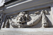 Antique, time-worn, sculptural decoration on the sunlit facade of a building, architectural detail, mascaron with a lion's head, and floral ornament elements with a ribbon.