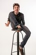 Expressive young adult sticking out tongue in a teasing gesture while sitting on a bar stool against a white background.