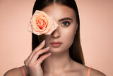 Fototapeta Tęcza - Beauty studio portrait of young beautiful woman holding rose against background in peach tones.